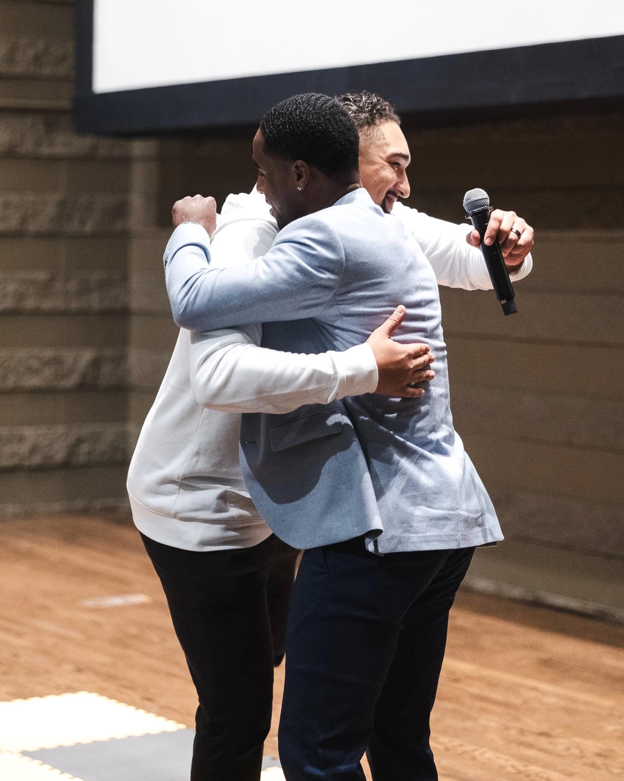 Two men hugging each other during a formal event, with one holding a microphone and smiling.