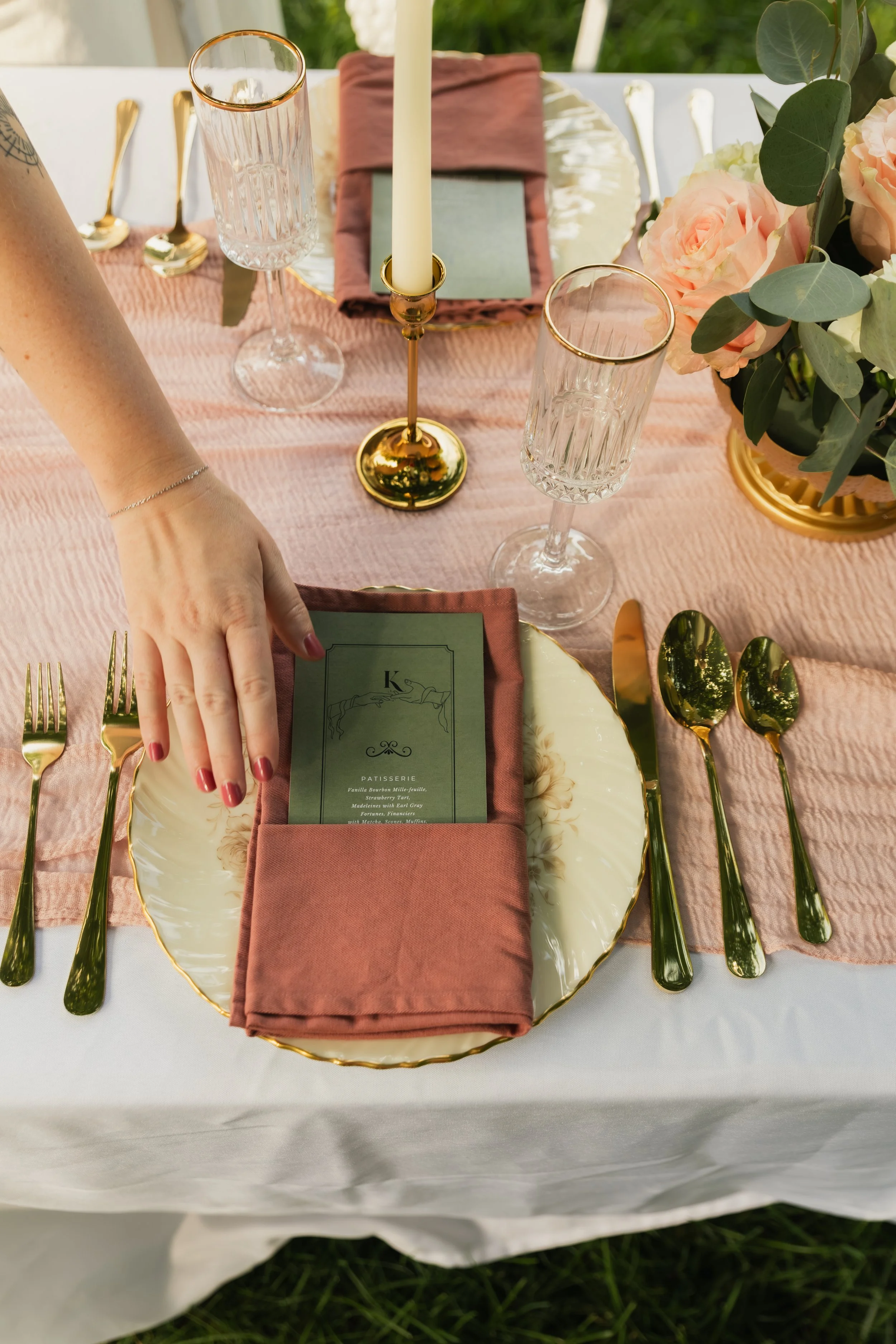 Elegant table setting with a pink tablecloth, gold cutlery, crystal glasses, a gold candleholder with a candle, a menu card, and a floral centerpiece of pink roses and greenery.