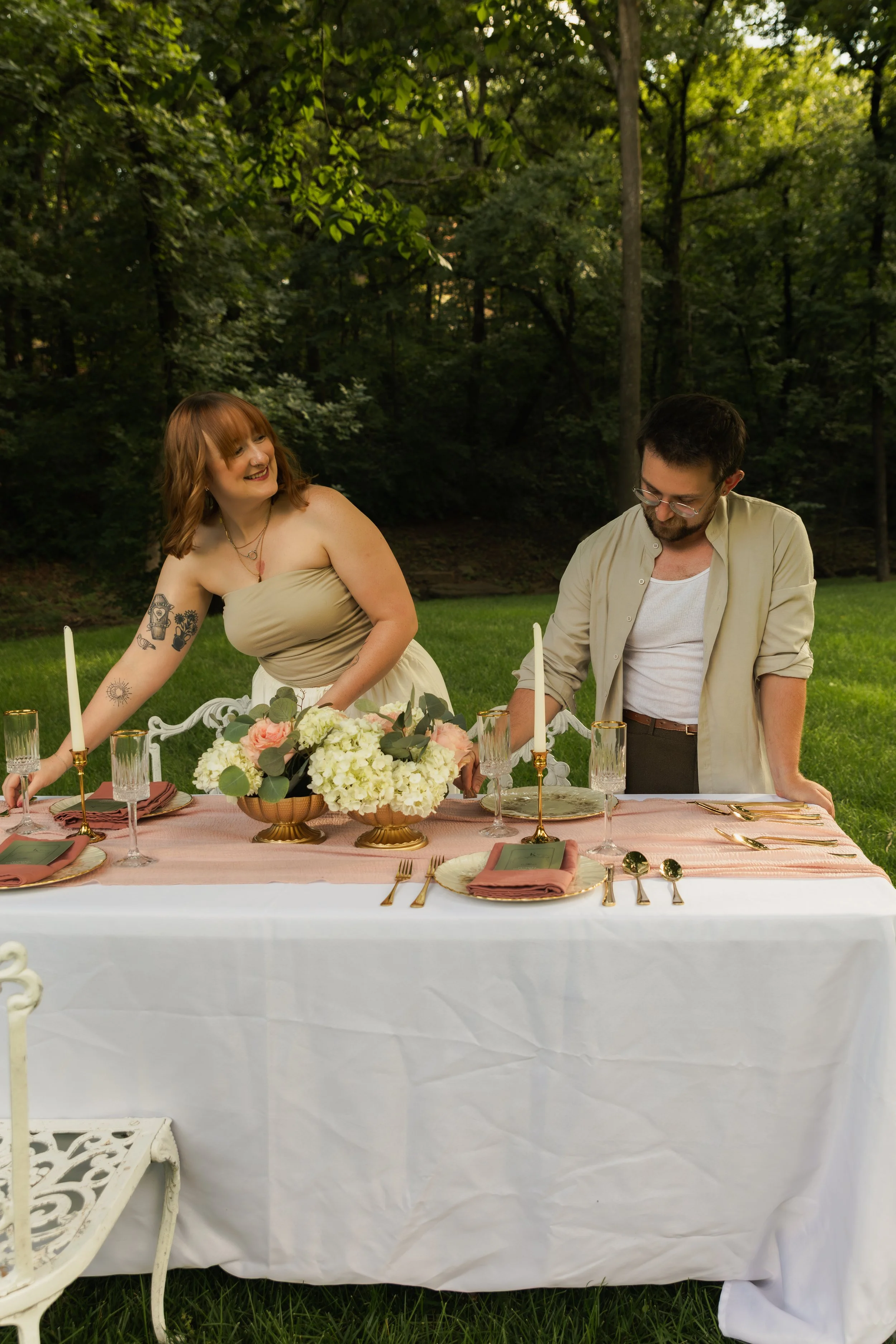 A couple is setting a decorative table outdoors in a green, wooded area. The woman has red hair, tattoos, and is smiling at the man. The man is wearing glasses and a light shirt. The table is decorated with a floral centerpiece, pink napkins, gold utensils, and candles.