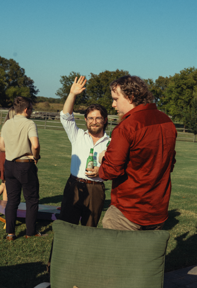 Two men standing outdoors on a sunny day talking, one with a beer bottle raising hand, other listening, in a park with trees and a clear sky.