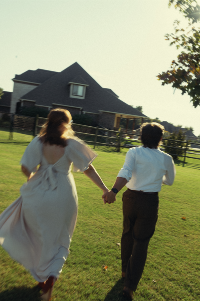 A woman and a man holding hands, running across a grassy yard near a house, during late afternoon or early evening.