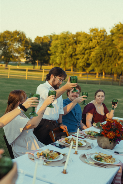 Group of four adults raising glasses at an outdoor dinner table with food and flowers during sunset.