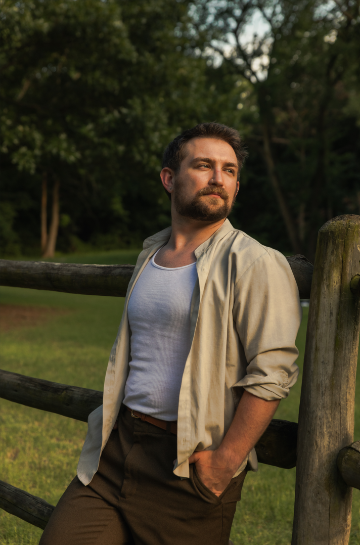 A man with a beard and short dark hair leaning against a wooden fence, looking off into the distance with a contemplative expression during sunset in a park with green trees.