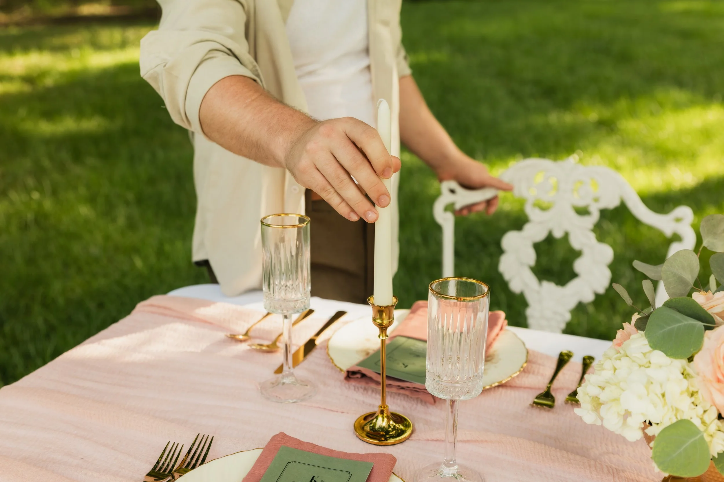 A person setting a table with elegant glassware and flowers outdoors.