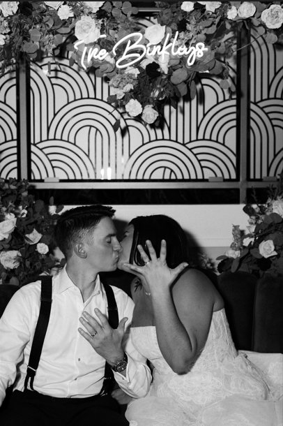 A couple leaning in for a kiss in a decorated setting with a floral arrangement and a neon sign that reads 'The Bunkers' in the background.
