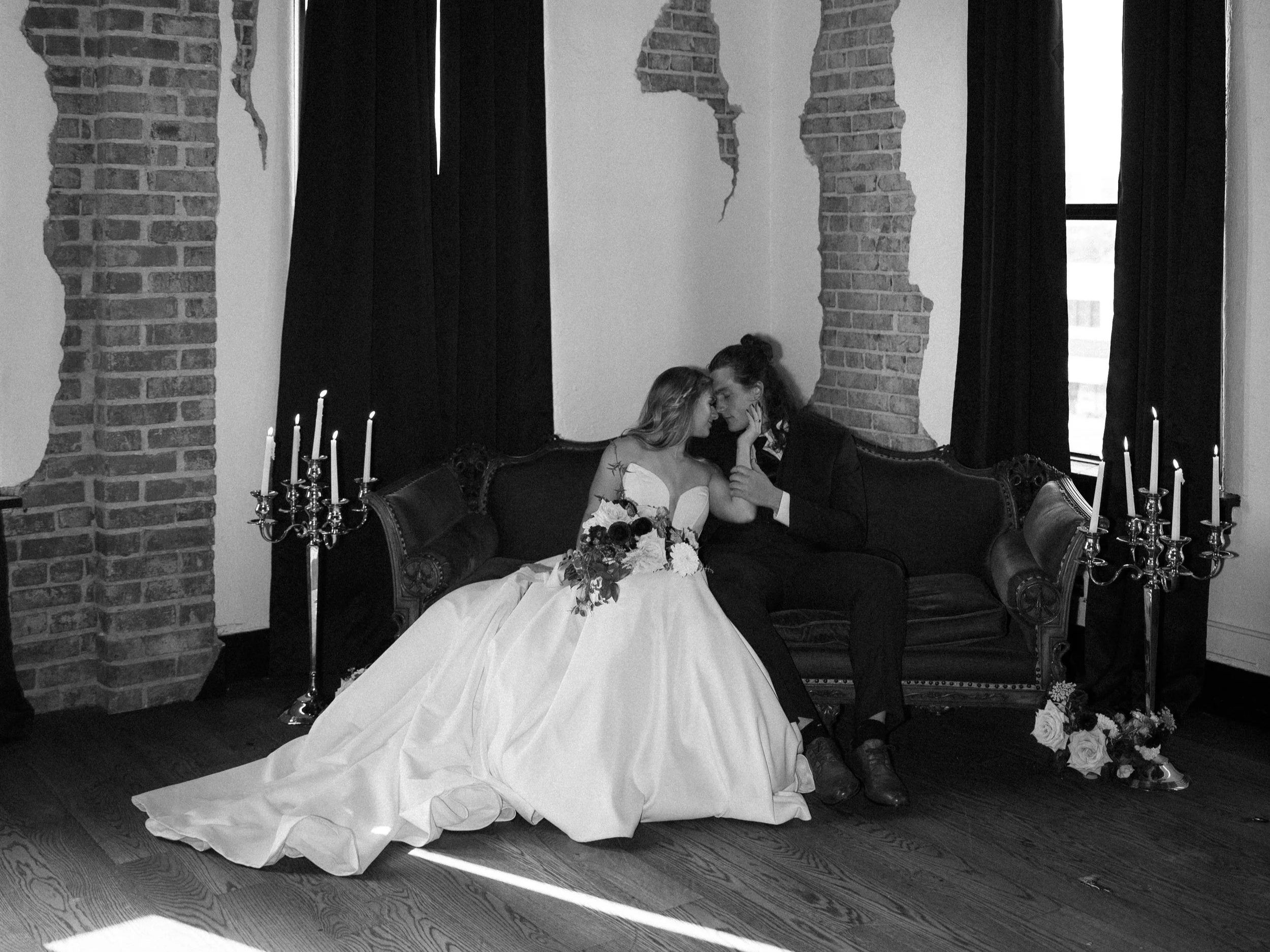 A bride in a wedding dress sitting on a vintage couch, holding a bouquet of flowers, with a groom in dark clothing gently touching her face. The scene is set indoors with brick walls, black curtains, and candelabras, creating a romantic atmosphere.