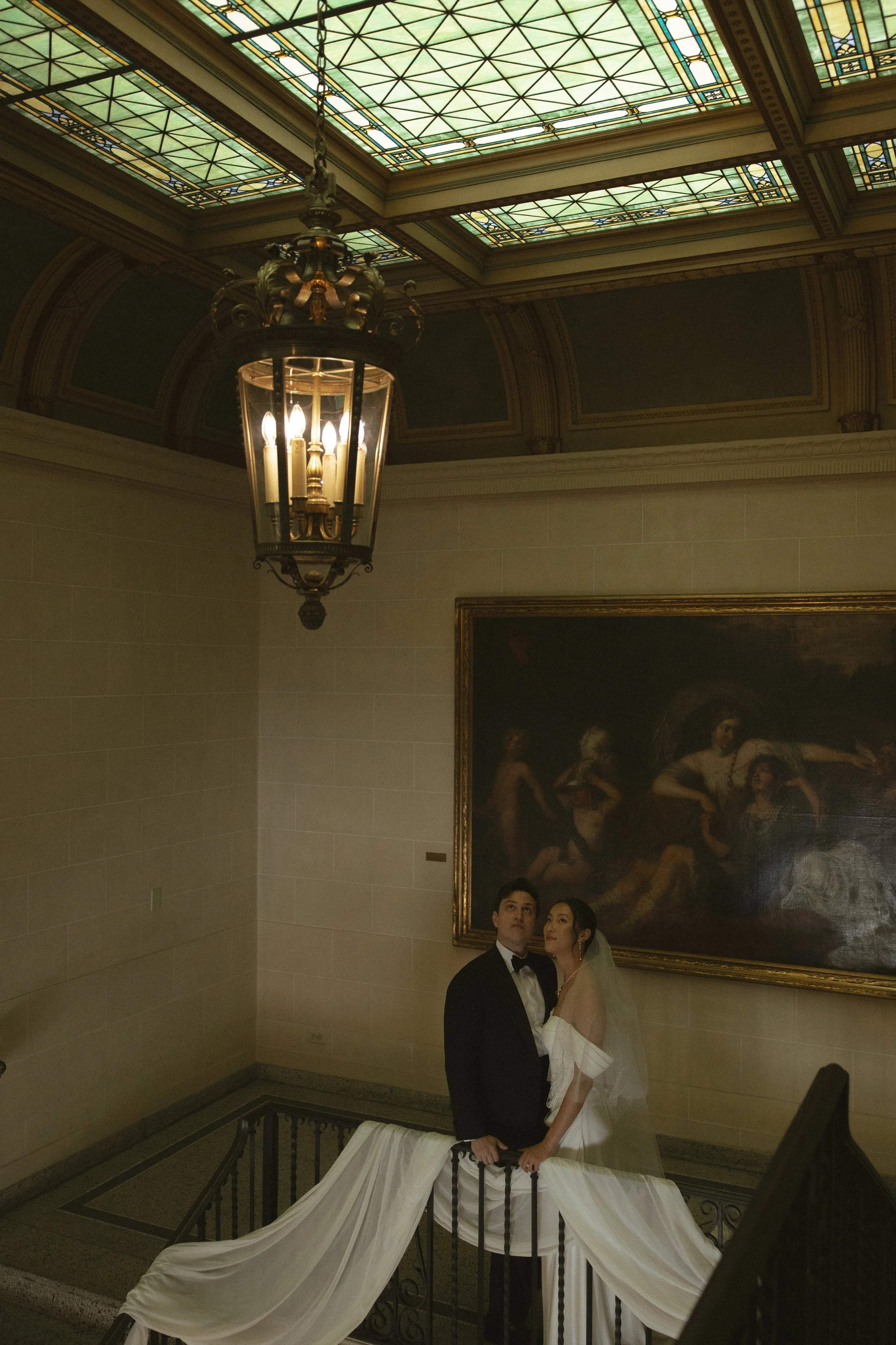 A bride and groom in wedding attire standing on a staircase in an elegant indoor space with stained glass ceiling and large oil painting. The bride is wearing a white dress with a veil, and the groom is in a tuxedo.
