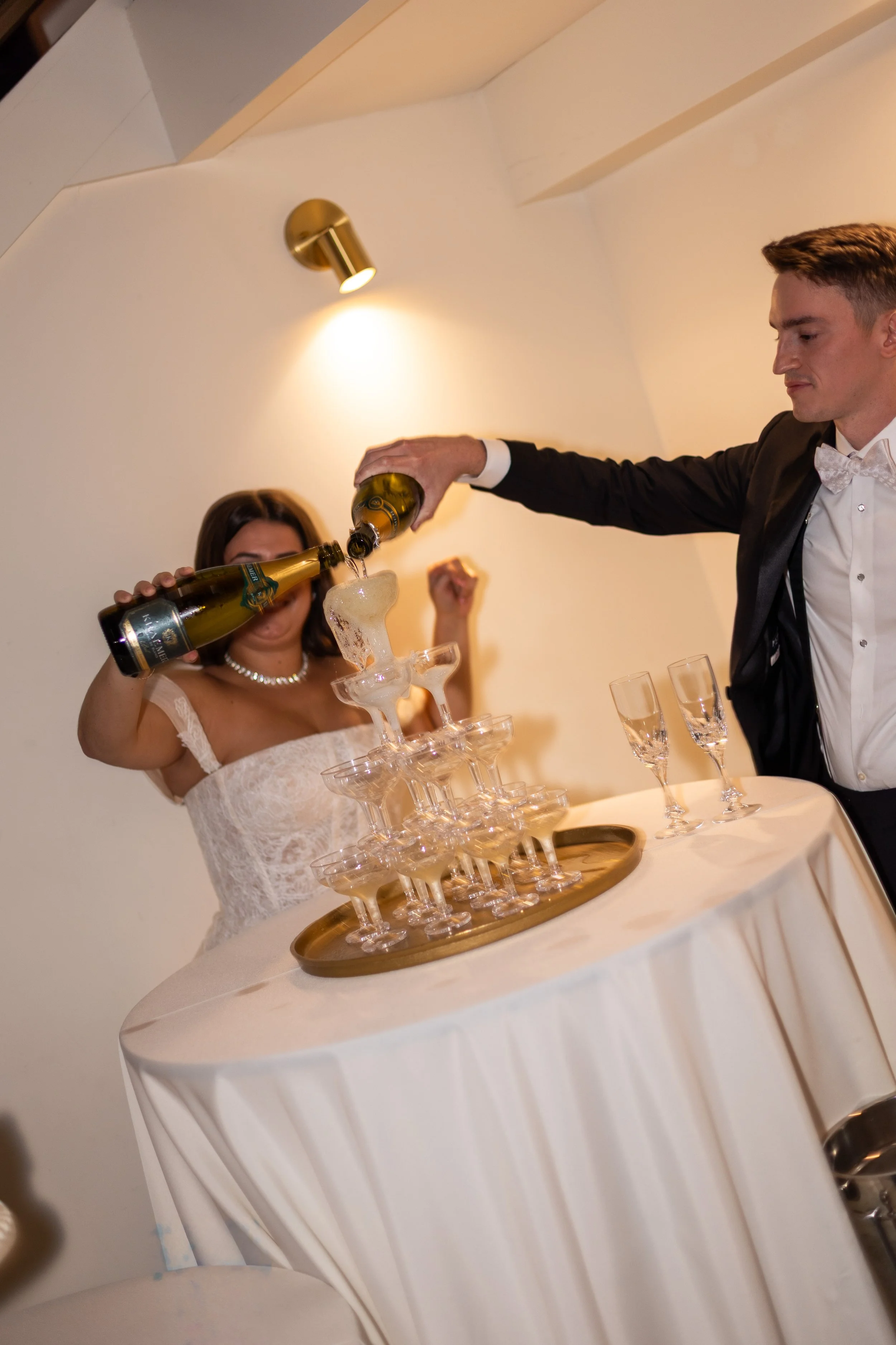 A woman in a white dress is pouring champagne into a glass from a bottle, standing next to a champagne tower on a table with a white tablecloth. A man in a black tuxedo with a bow tie is standing nearby, and there are empty champagne glasses on the table.
