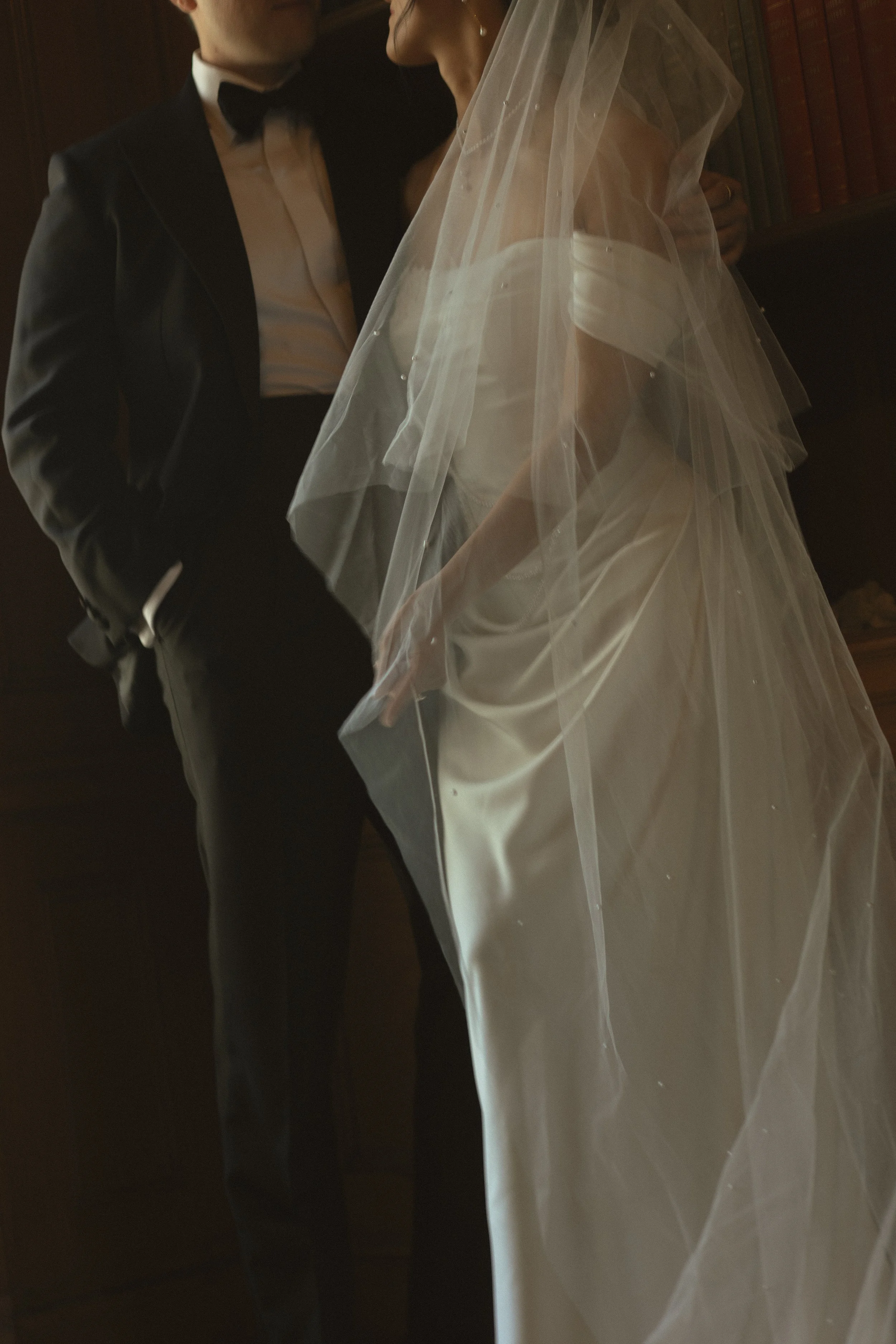 A couple in formal wedding attire is standing close together indoors, with the bride wearing a white wedding gown and veil, and the groom in a black tuxedo and bow tie.