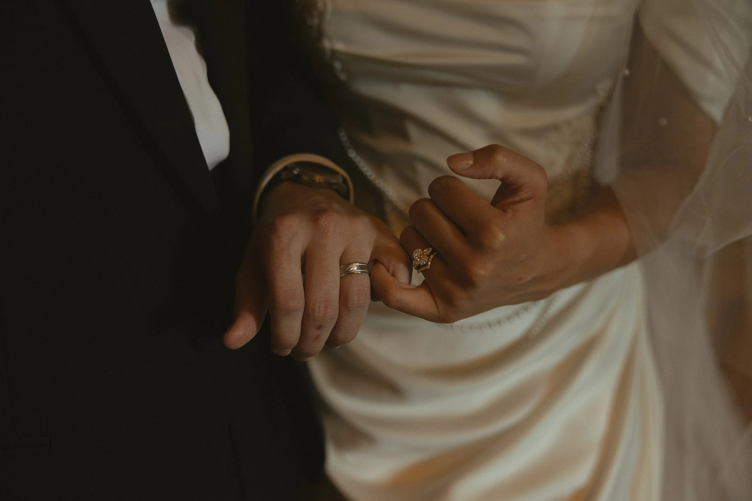 Close-up of a couple holding hands, showing wedding rings, with the bride wearing a white dress and the groom in a dark suit.