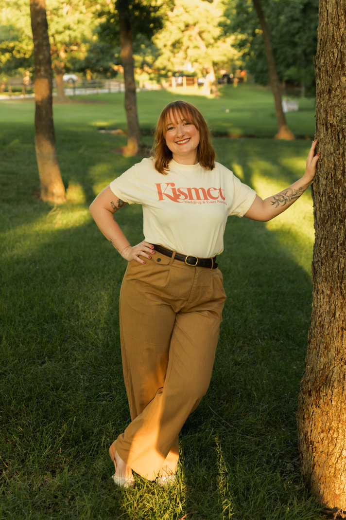 A woman with reddish-brown hair, wearing a white T-shirt with 'Kismet' printed on it, tan pants, and a black belt, standing outdoors in a park with trees and green grass, leaning against a tree, smiling at the camera.