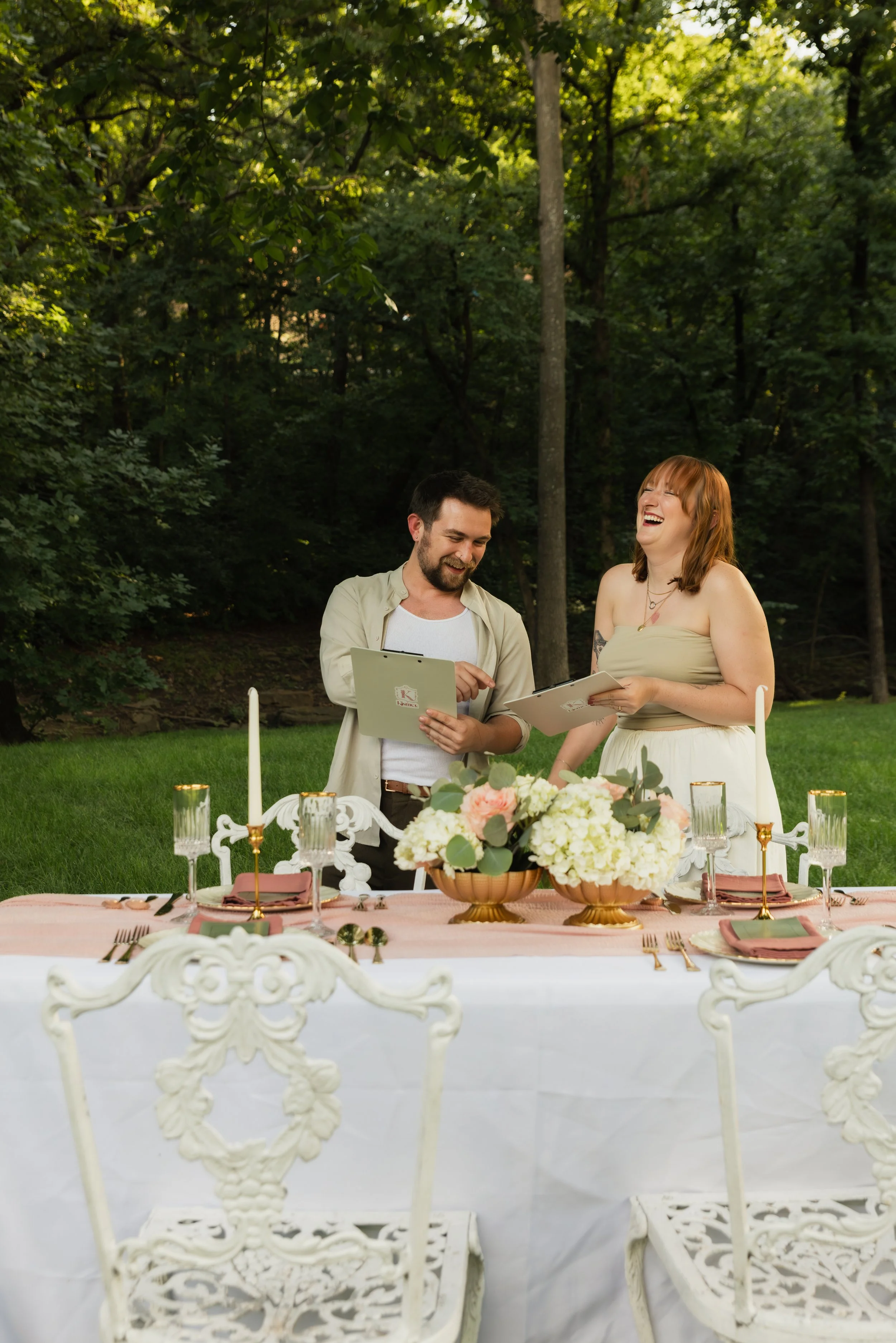 Two people, a man and a woman, are standing outdoors at a decorated table, smiling and looking at each other. The table is set with glassware, napkins, and a floral centerpiece, indicating a celebration or special event. The background features lush green trees and foliage.