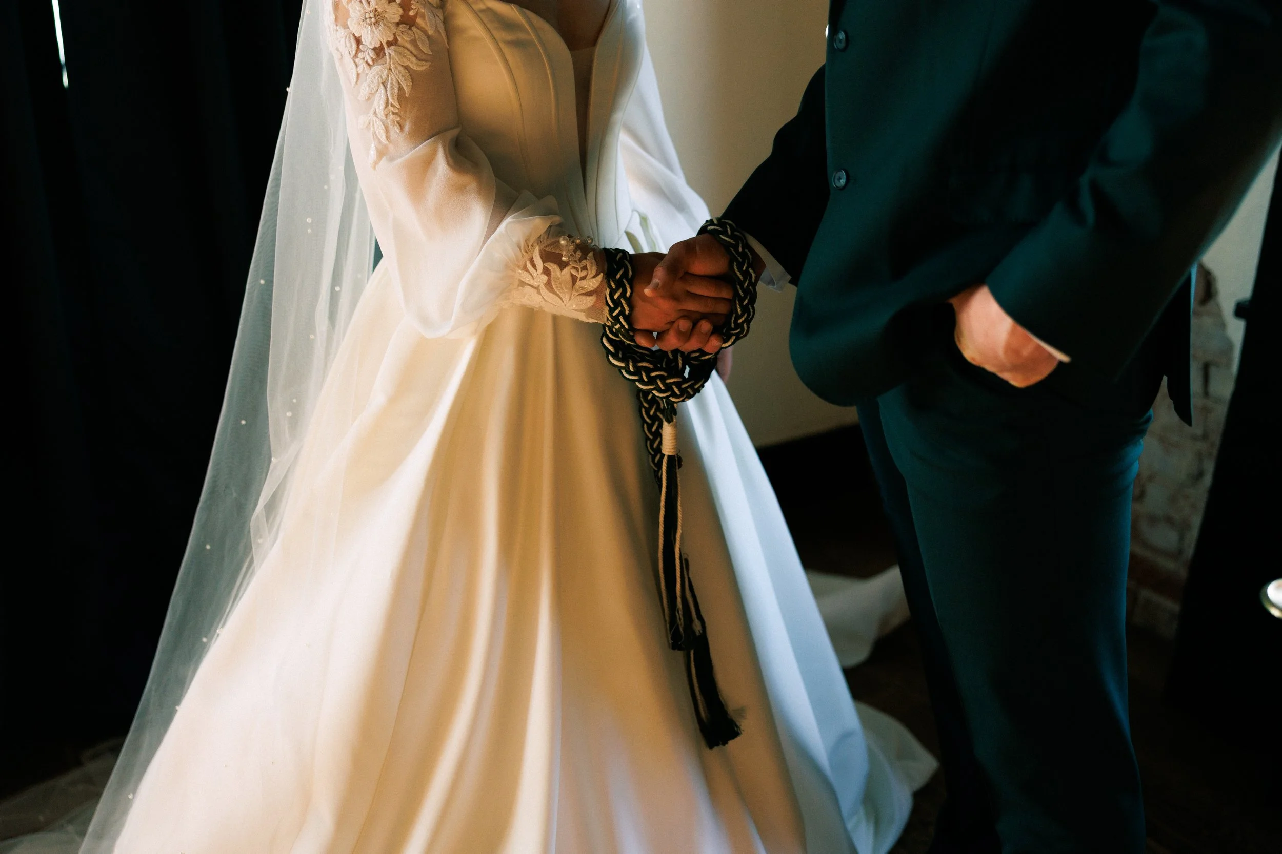 Close-up of two people holding hands, one wearing a wedding dress with floral embroidery on the sleeve and the other in a dark suit, during a wedding ceremony.