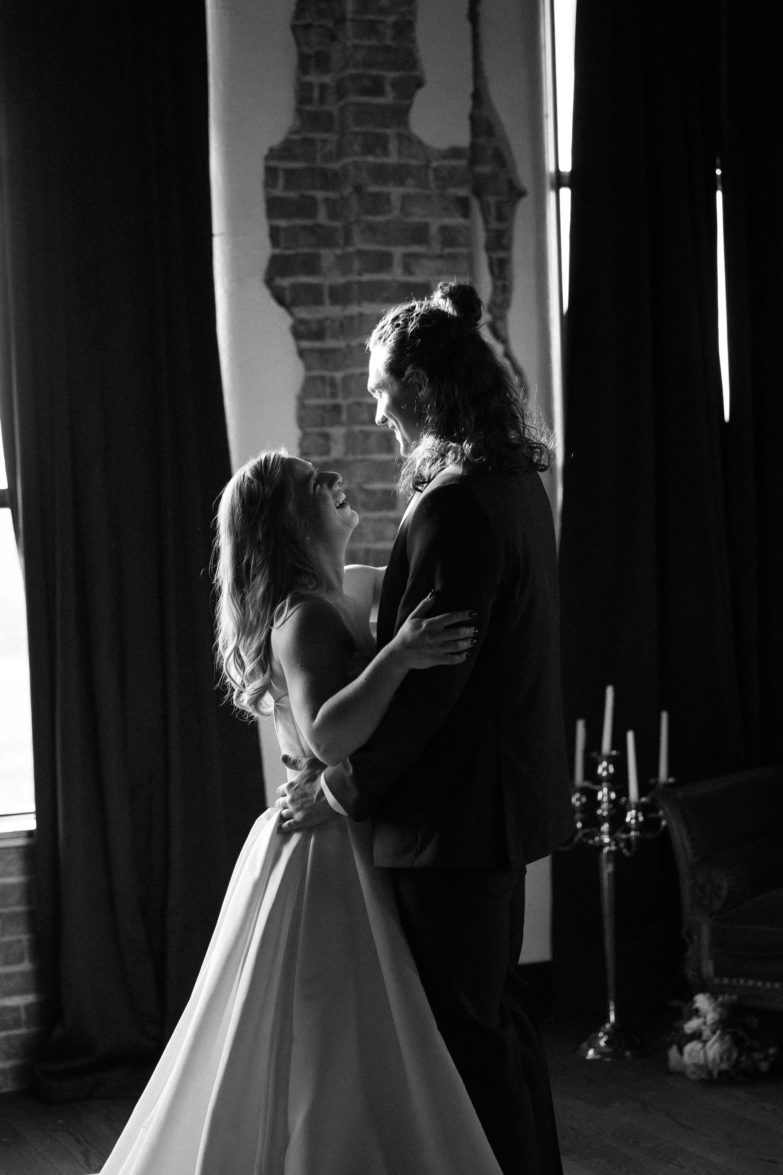 A black and white photo of a couple dancing indoors, with a brick wall and a window with curtains in the background.