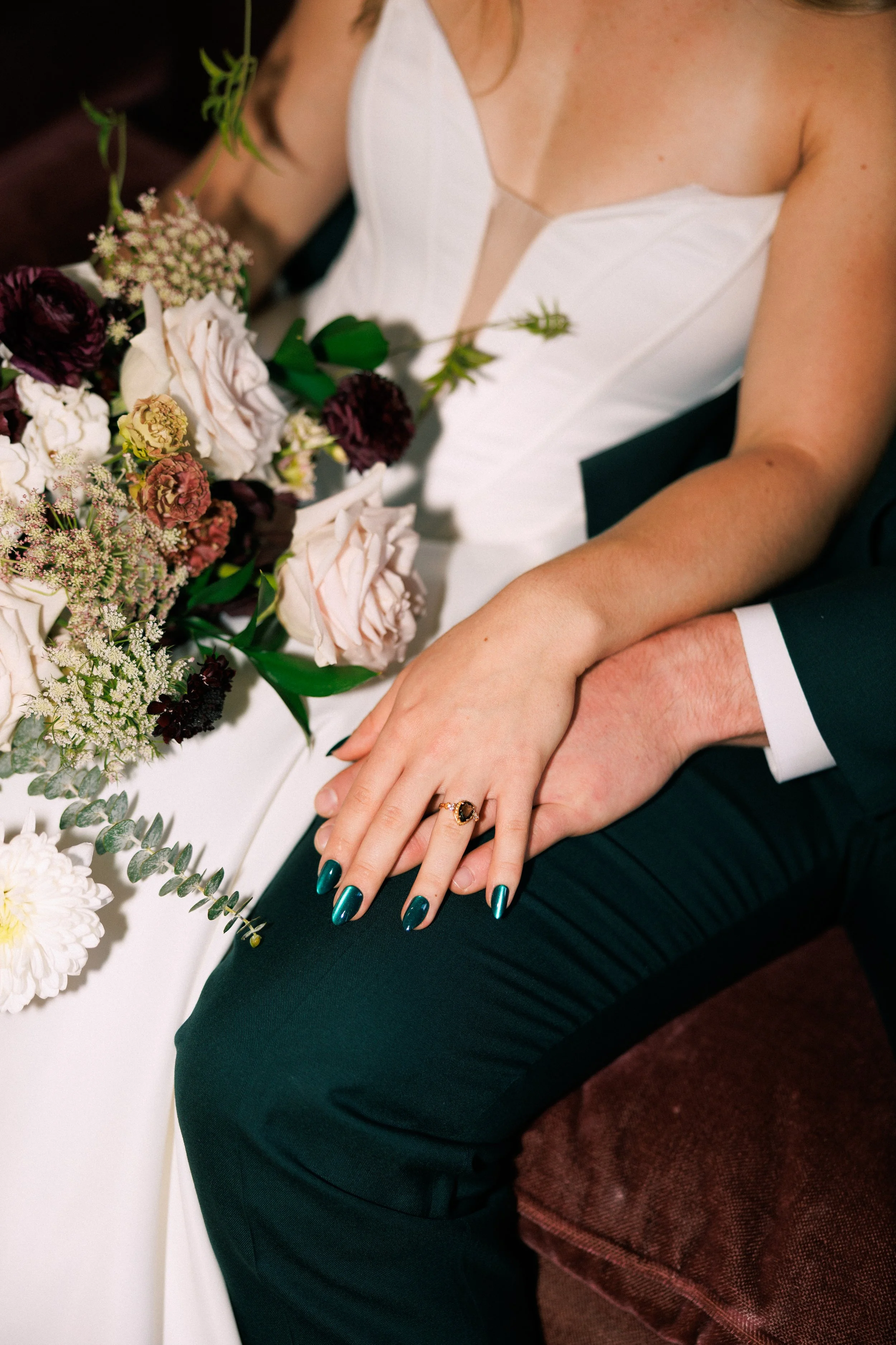 Wedding couple holding hands, with the woman's hand displaying a ring, near a bouquet of flowers on a white wedding table. The woman is wearing a white dress and teal nail polish, and the man is in a dark suit.