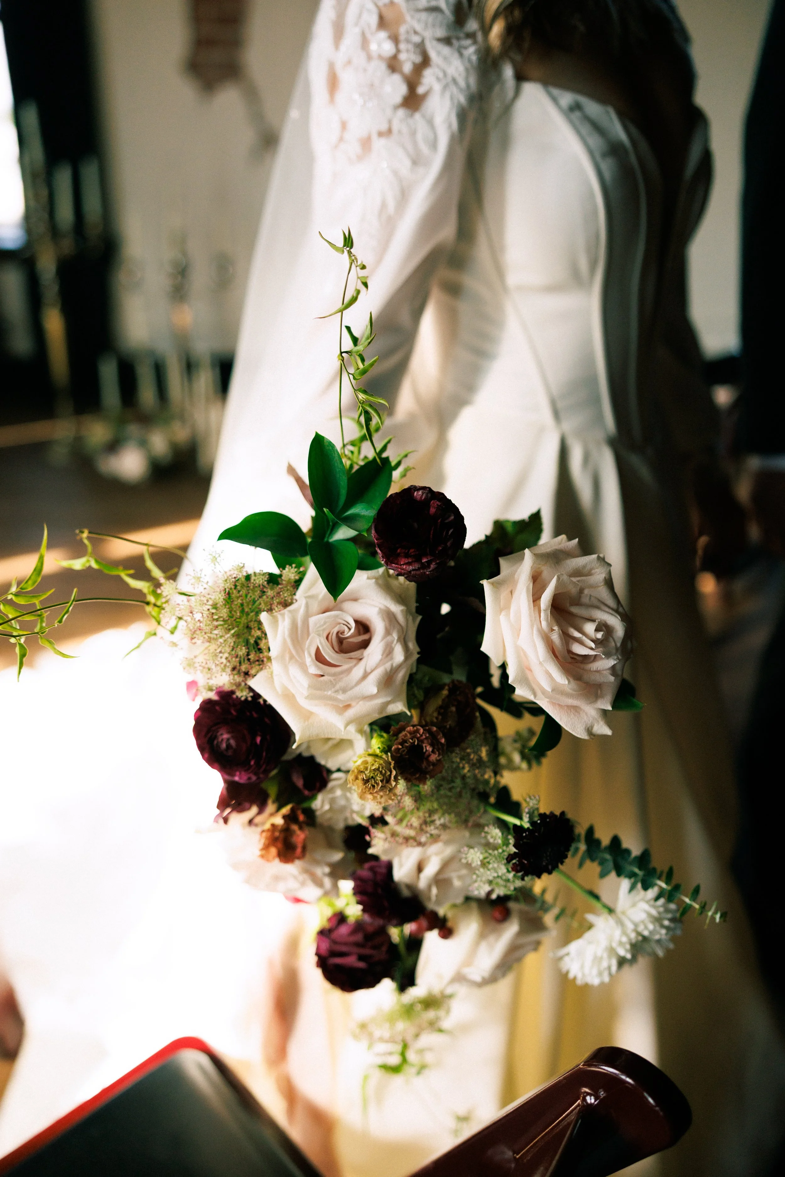 Close-up of a wedding bouquet with white roses, dark purple flowers, green leaves, and small white flowers, resting on a white surface, with a blurred background.