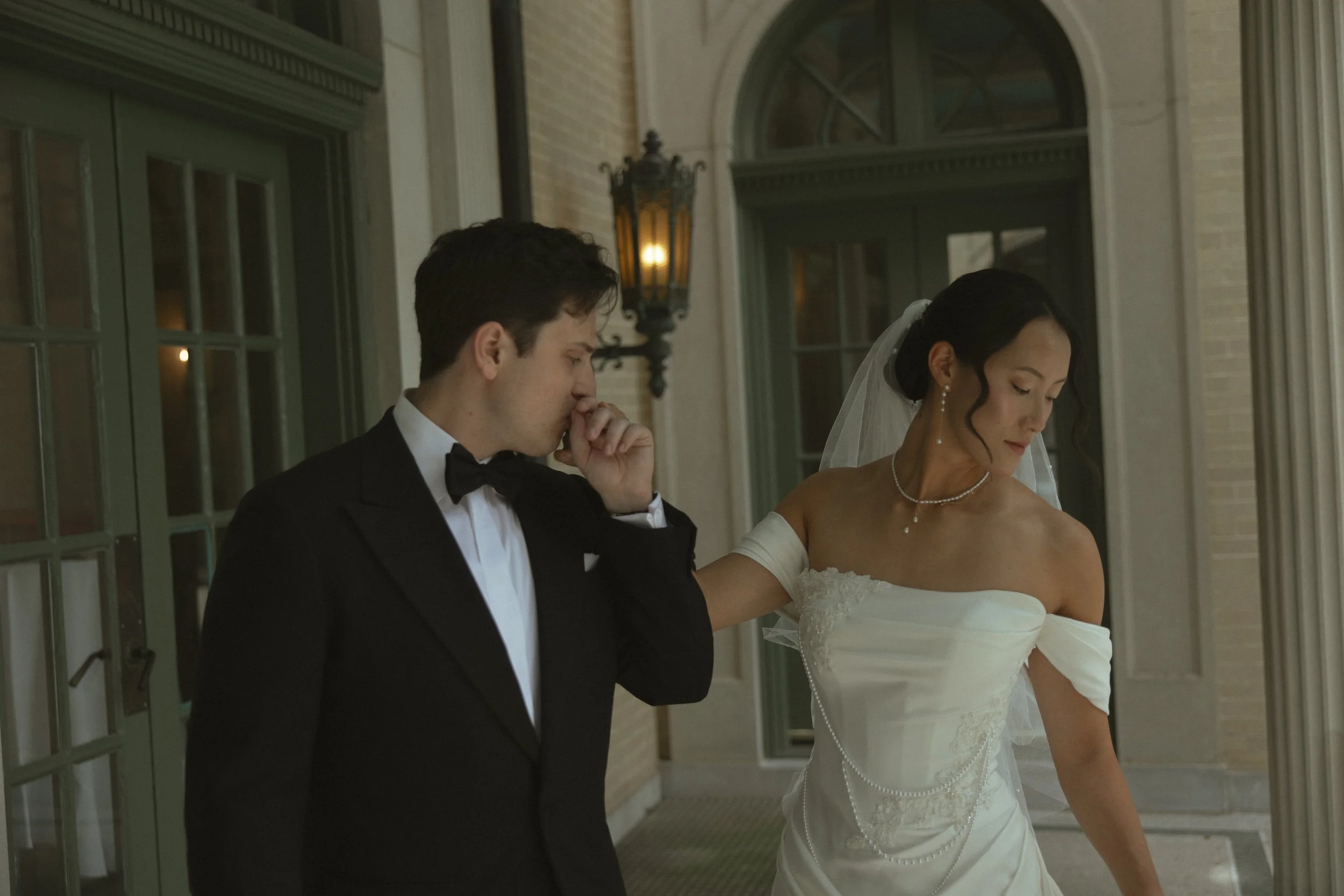 A bride and groom standing outside on a porch at night, with the groom in a tuxedo and the bride in a white wedding dress, both with solemn expressions.