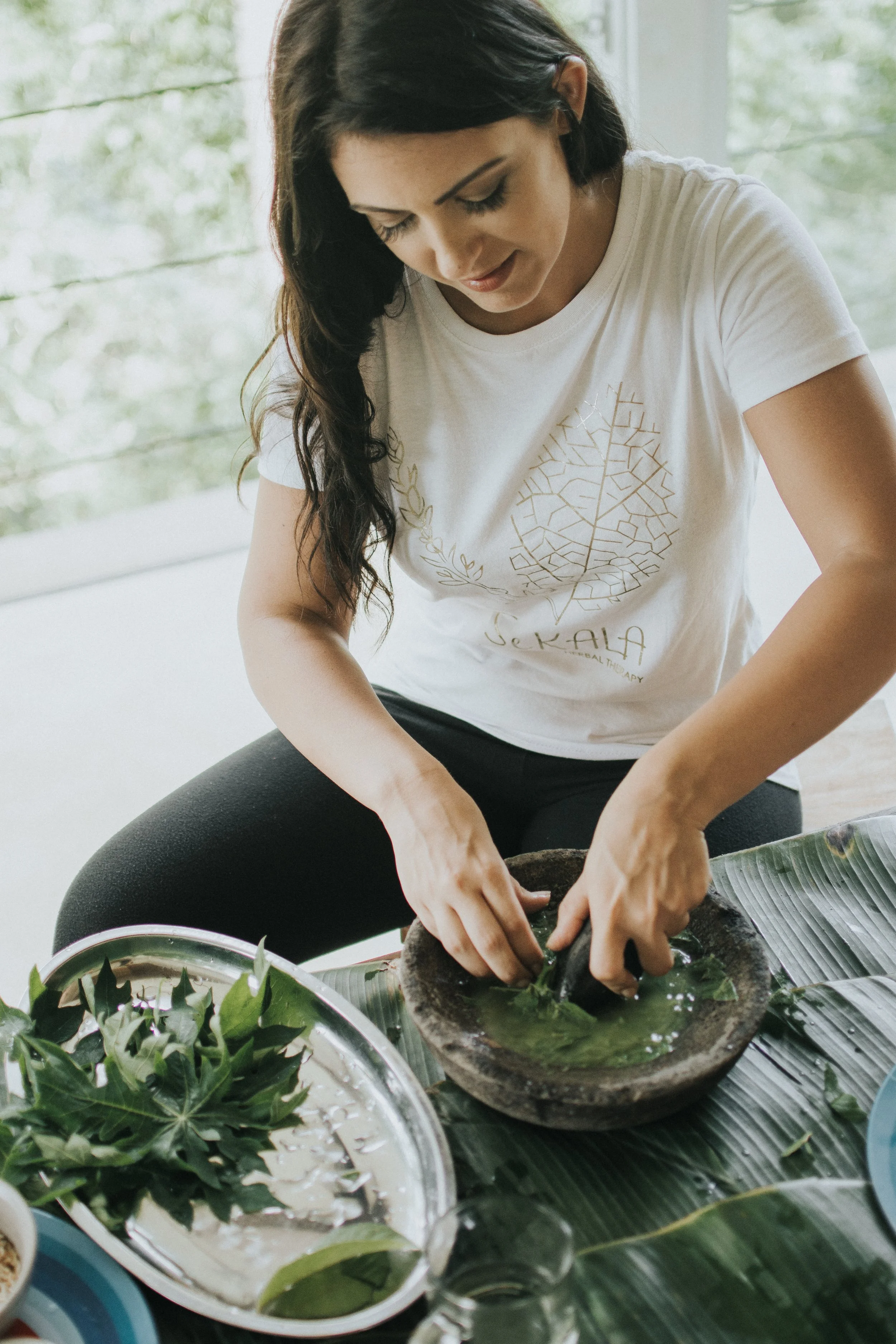 A woman in a white T-shirt with a leaf design is making traditional herbal medicine with green leaves and herbs, using a stone mortar and pestle near a window with green trees outside.