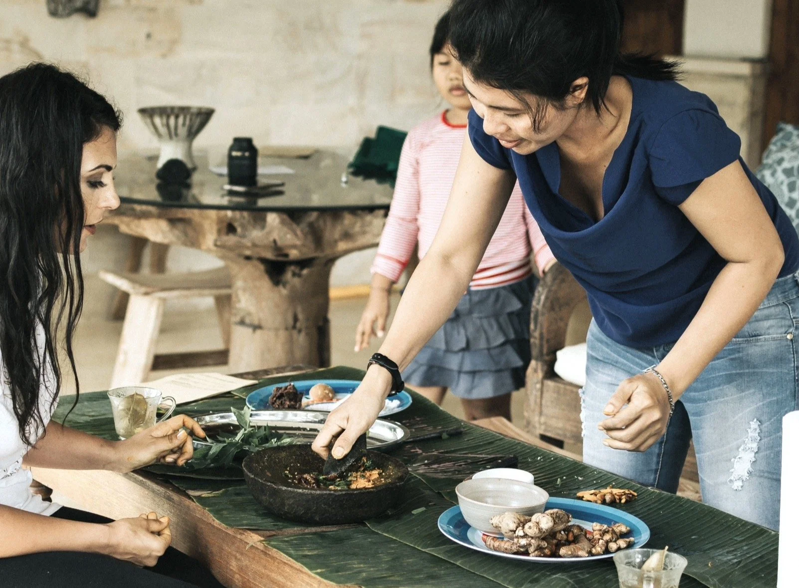 Two women and a girl at a dining table with food, one woman is serving food from a stone mortar, another woman is sitting, and the girl is standing in the background.