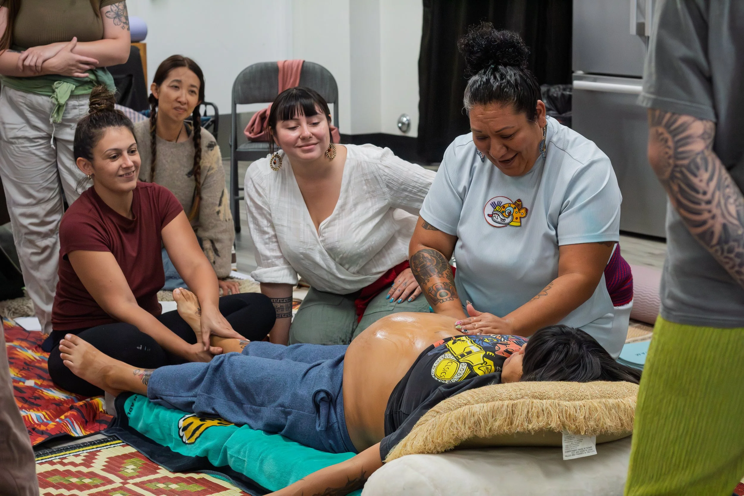 Group of women participating in a hands-on massage workshop, with one woman lying face down, receiving a massage from an instructor, and others observing and smiling.