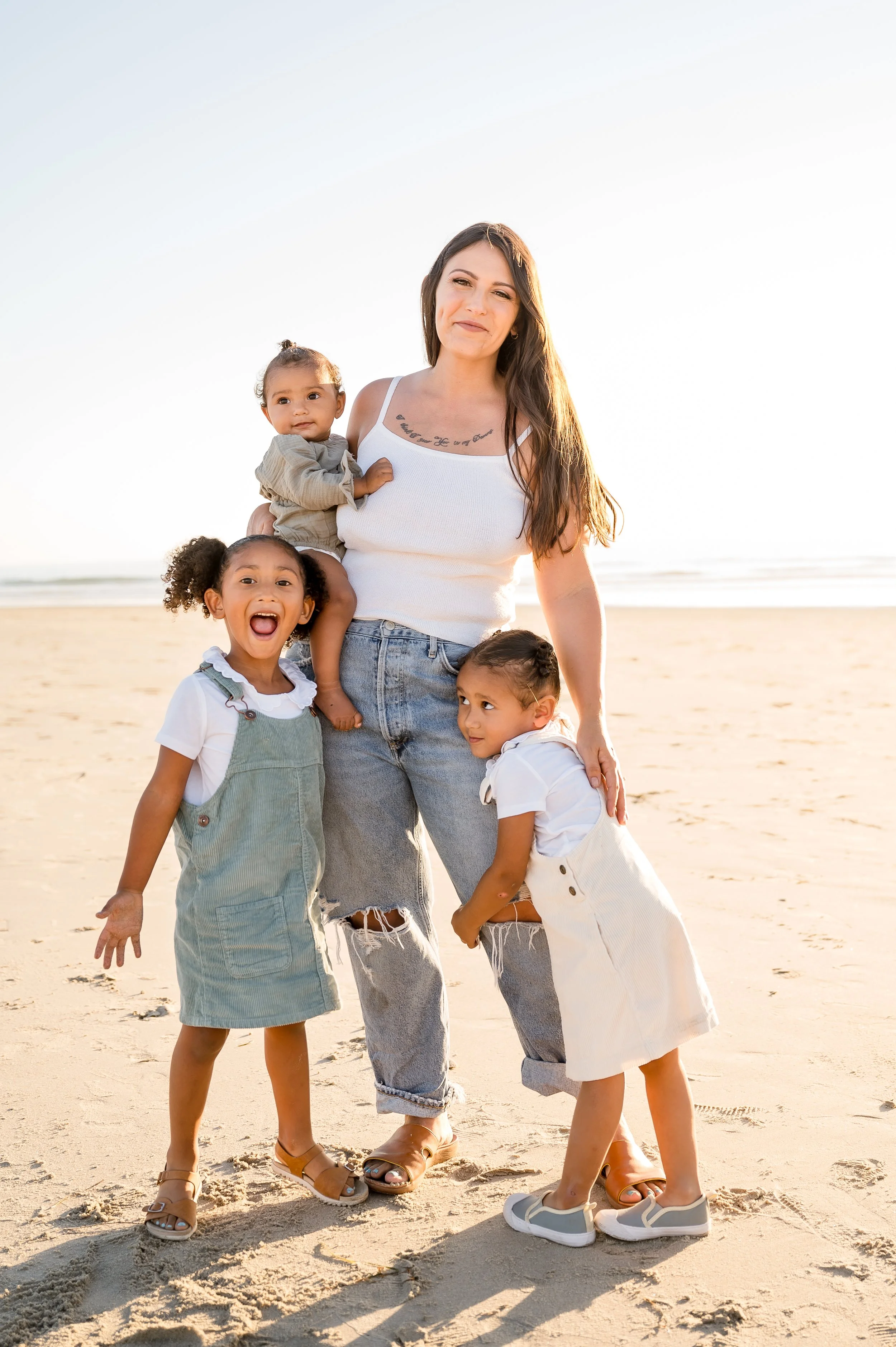 A woman with long brown hair standing on a sandy beach with four young girls, all smiling and enjoying a sunny day.