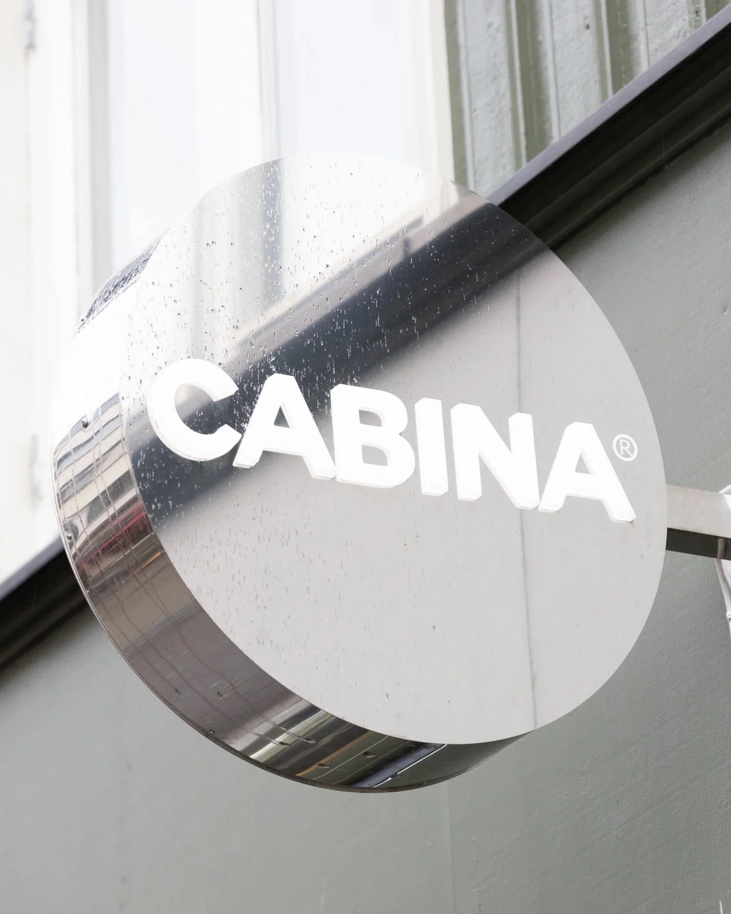 Refleksjon av utendørs bygning og blå himmel på et rundt firmaskilt med teksten 'CABINA'.