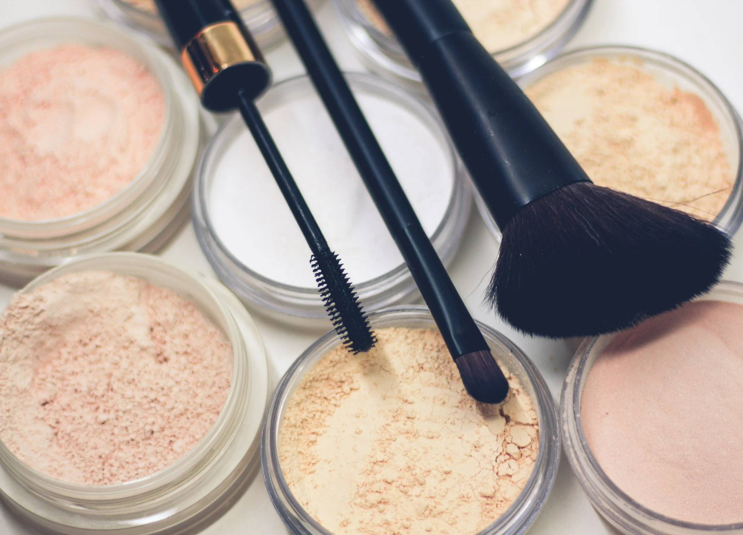 Bilde av flere åpne krukker med ulike fargede og teksturerte kosmetiske produkter, sammen med makeup-børster.