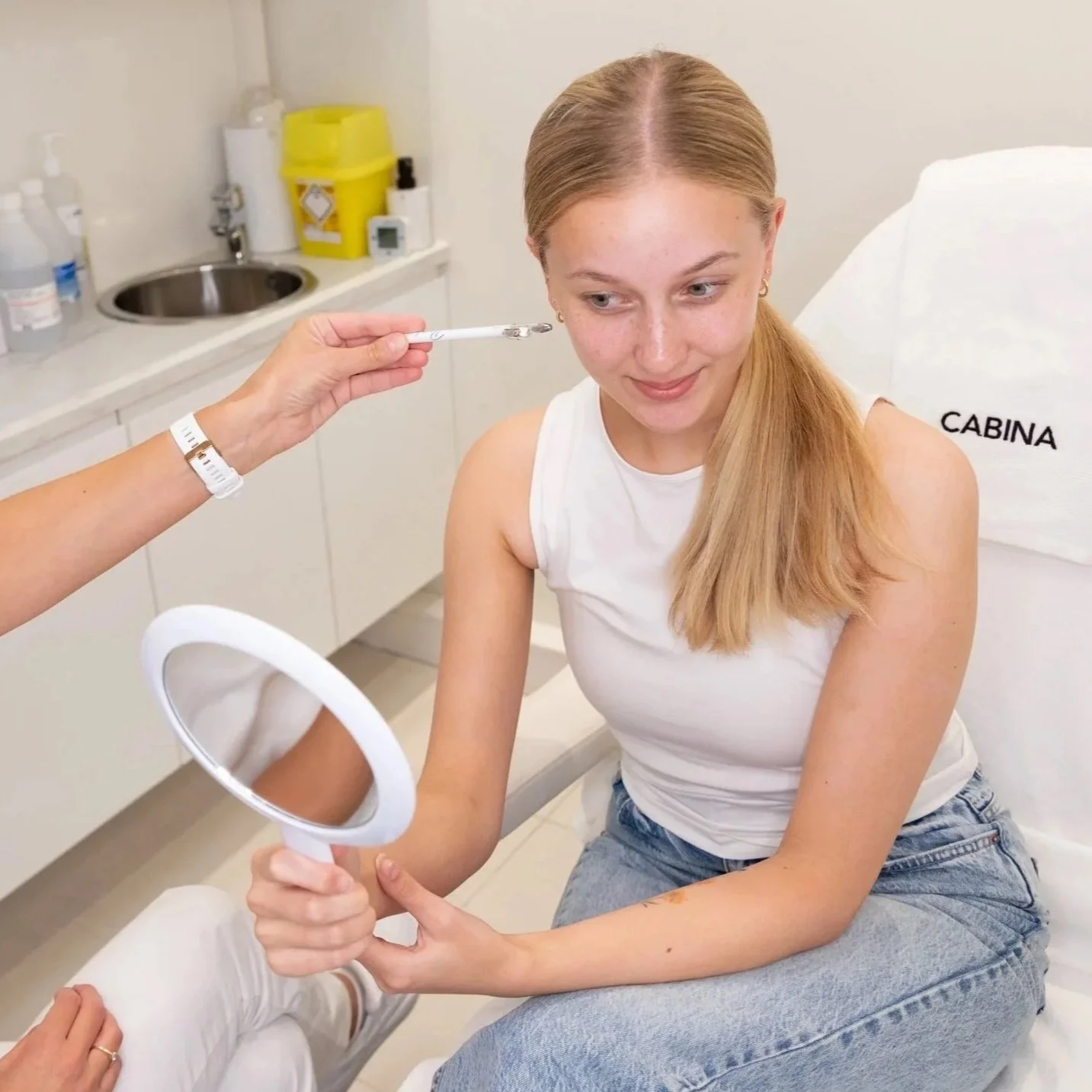 En ung kvinne med langt, lyst hår sitter i en klinikk og får gjennomført en hudanalyse.