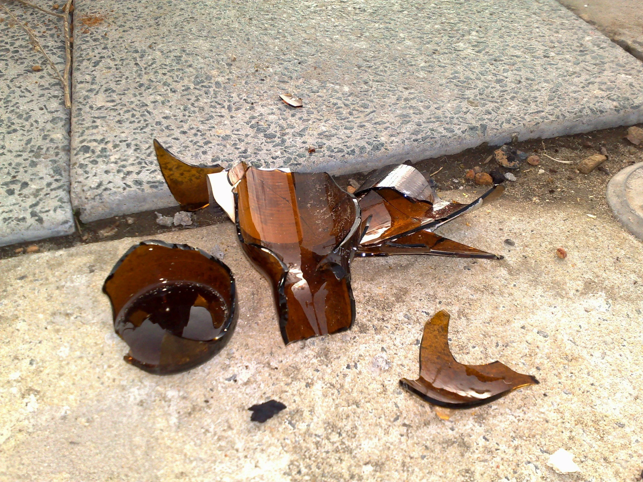 Broken brown glass bottles and pieces of shattered glass on a concrete sidewalk.