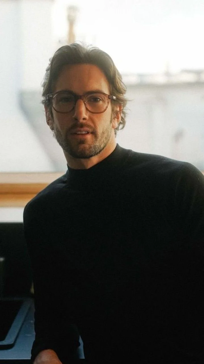 A man with wavy dark hair, wearing glasses and a black shirt, sitting indoors with a bright background.