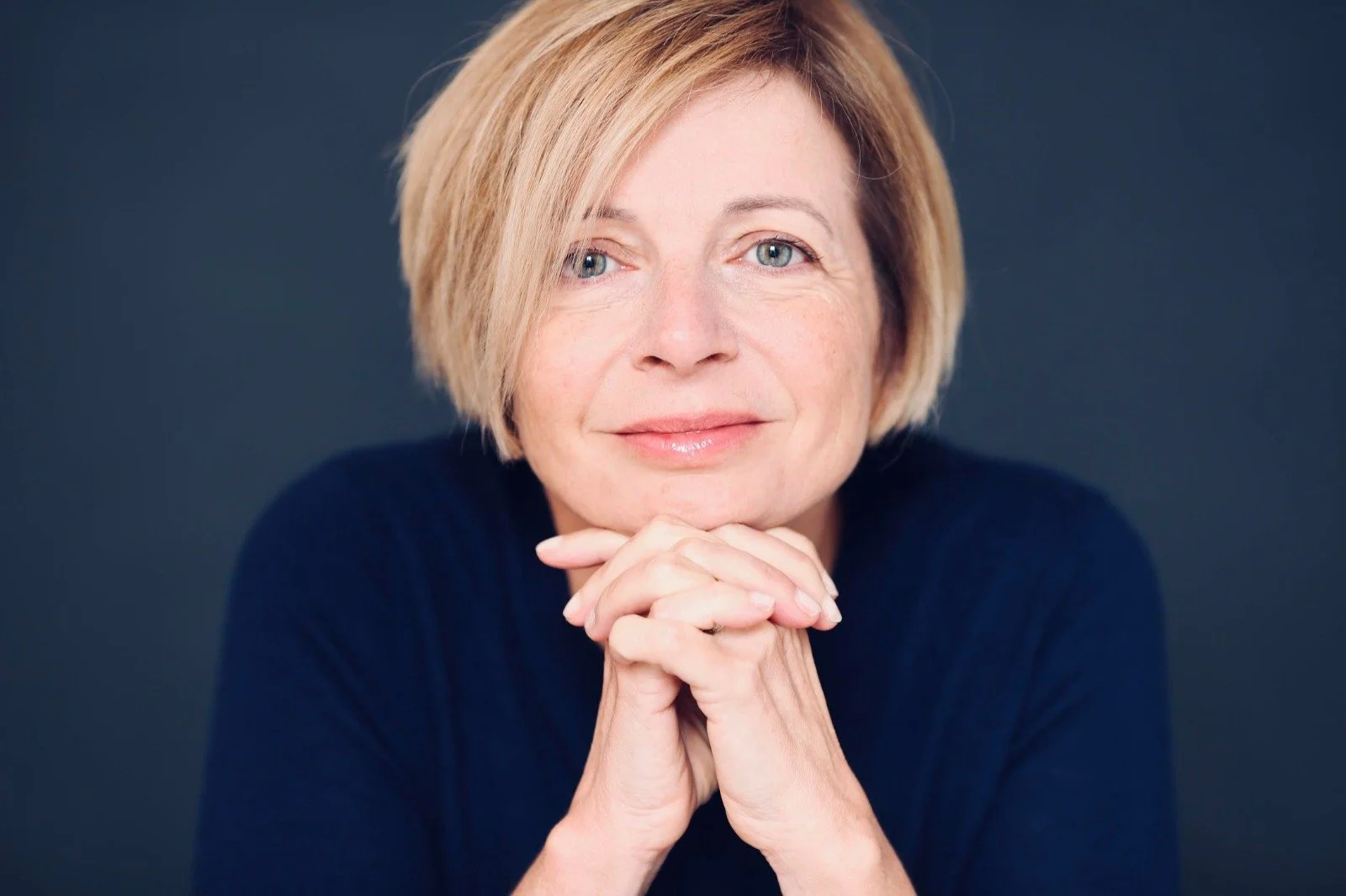 Close-up portrait of a middle-aged woman with short blonde hair, blue eyes, and a gentle smile, resting her chin on her clasped hands against a dark background.