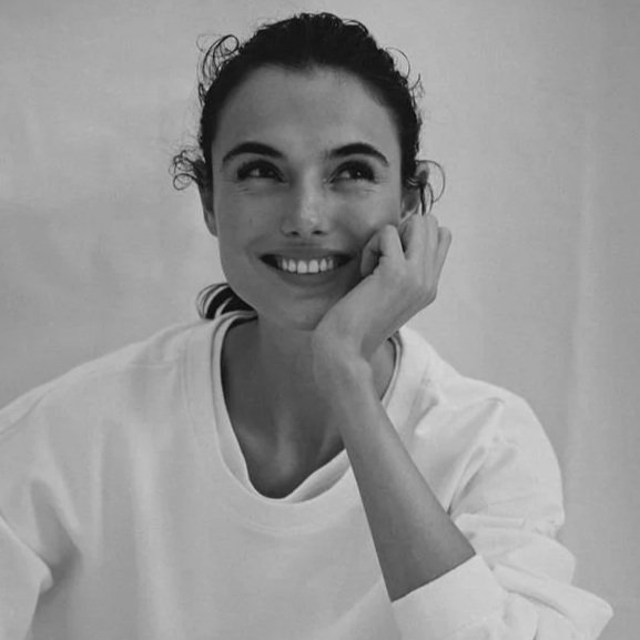 A woman with dark curly hair smiling and resting her face on her hand, wearing a white shirt.