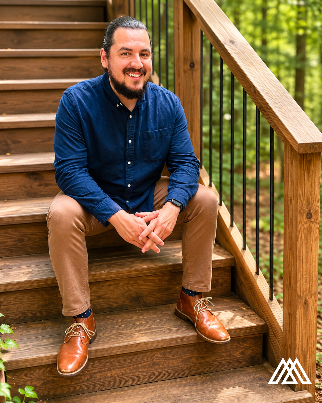 A man with dark hair, beard, and moustache seated on wooden stairs outdoors, wearing a navy blue shirt, tan pants, and brown shoes with black socks, smiling at the camera.