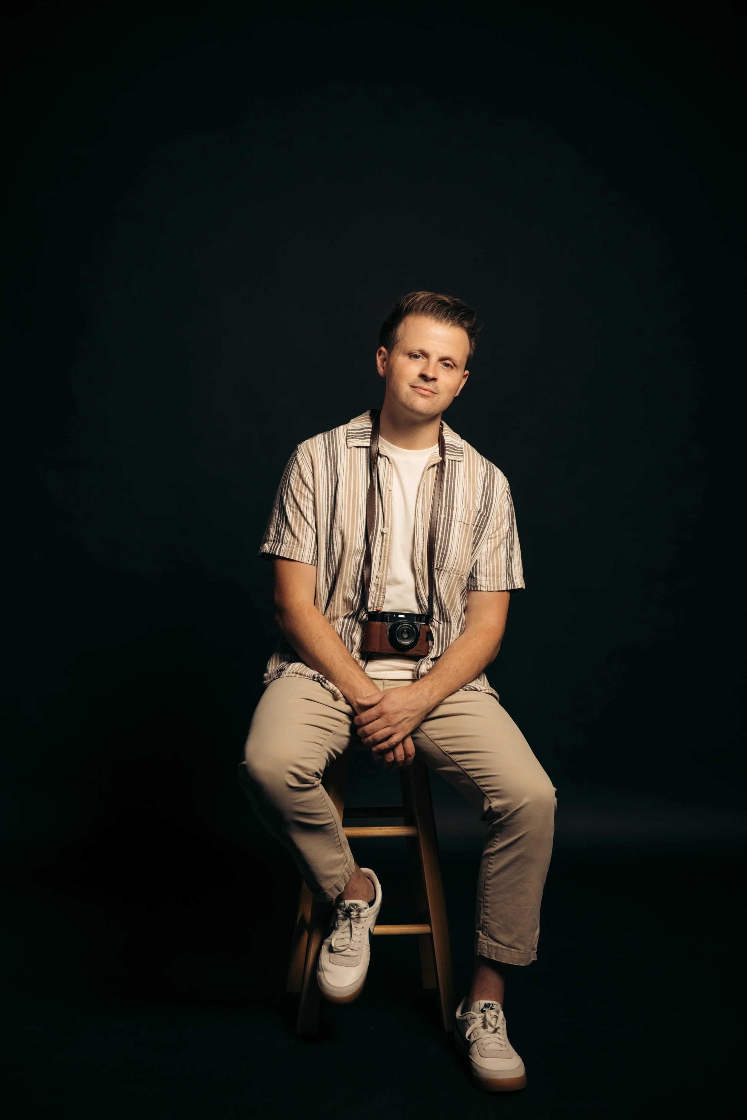 A young man wearing beige pants, white sneakers, a white t-shirt, and a striped short-sleeve shirt sitting on a wooden stool against a dark background. He has a camera hanging around his neck and is sitting with one leg crossed over the other, looking confidently at the camera.