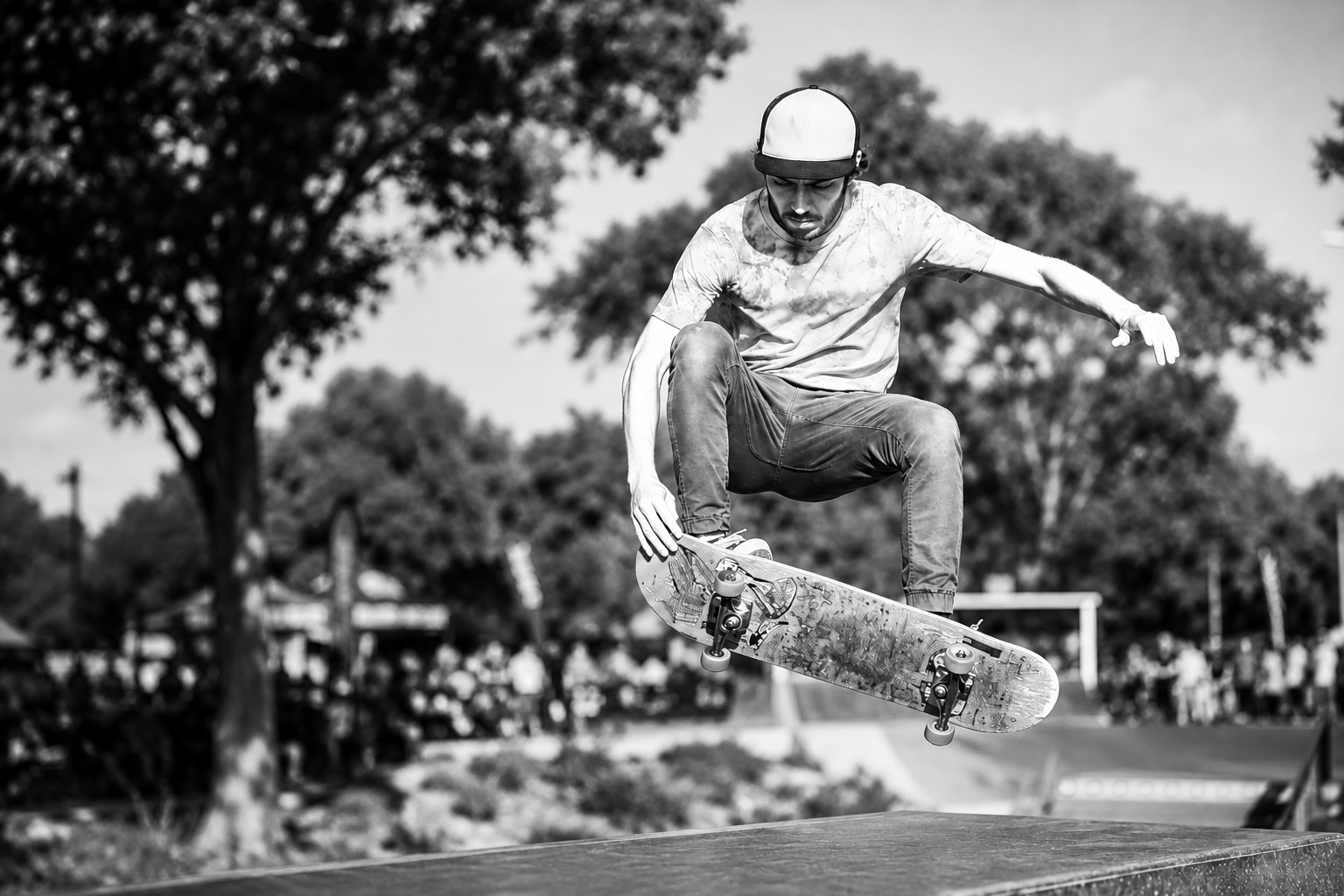 A skateboarder is mid-air performing a trick at a skatepark in a black and white photo, with trees and spectators in the background.