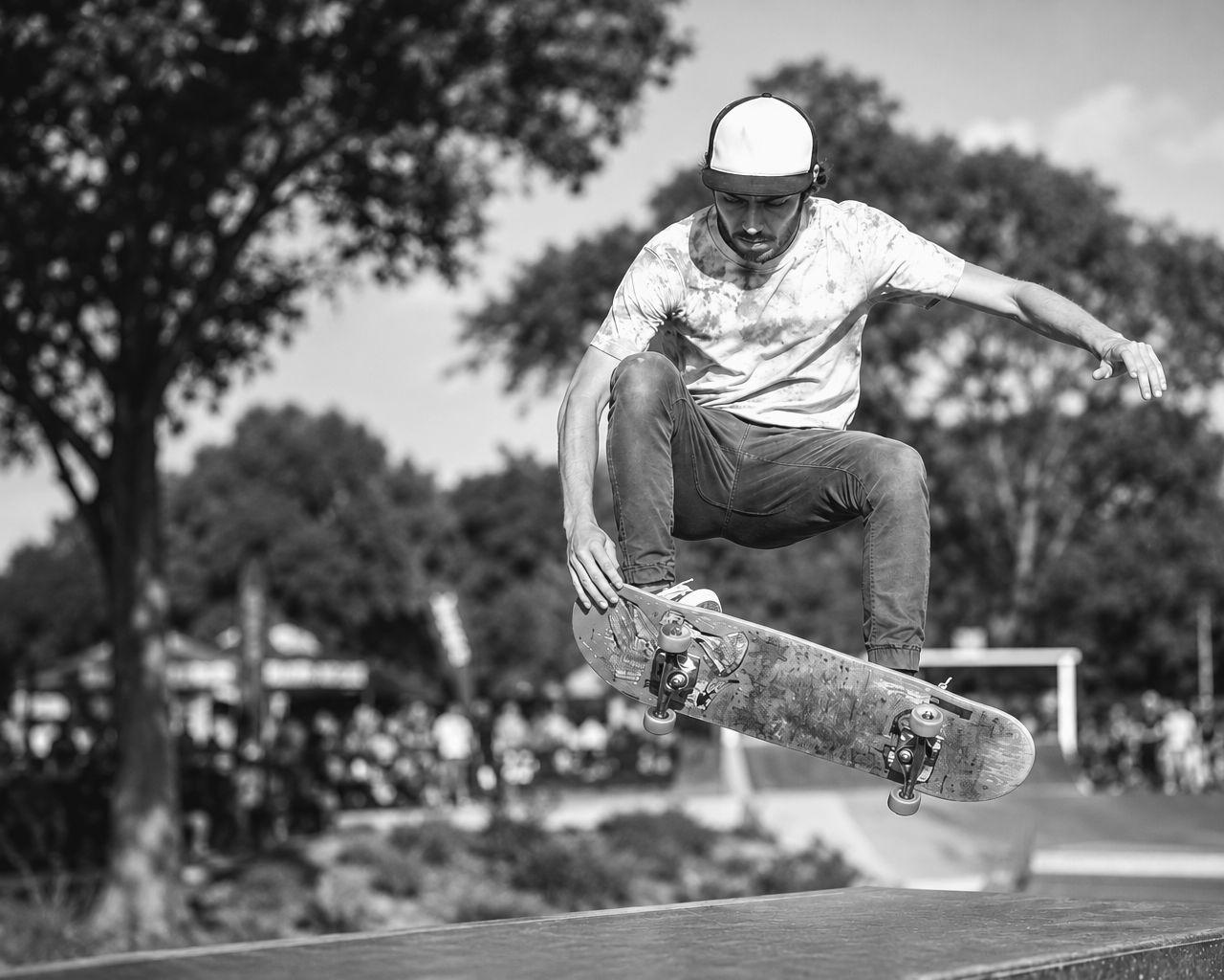 A skateboarder wearing a helmet performing a jump at a skatepark with trees and spectators in the background.