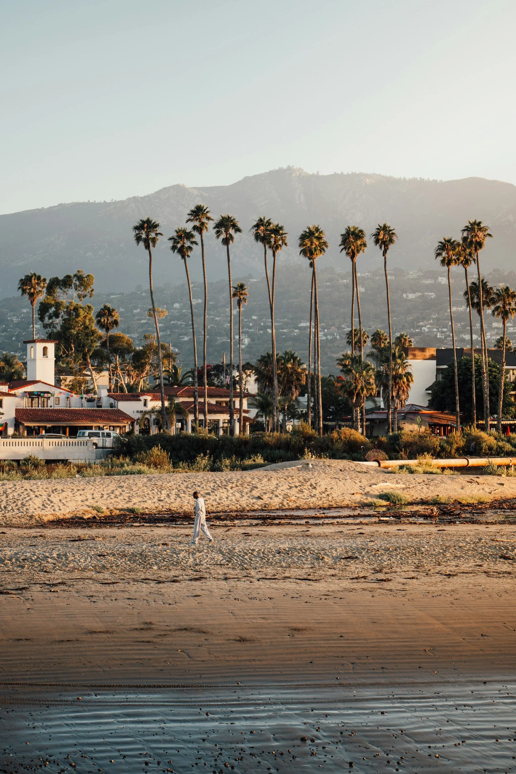 A person dressed in white walking along a beach with rippled wet sand, water, and scattered small shells or rocks, with houses, tall palm trees, and mountains in the background during sunset.