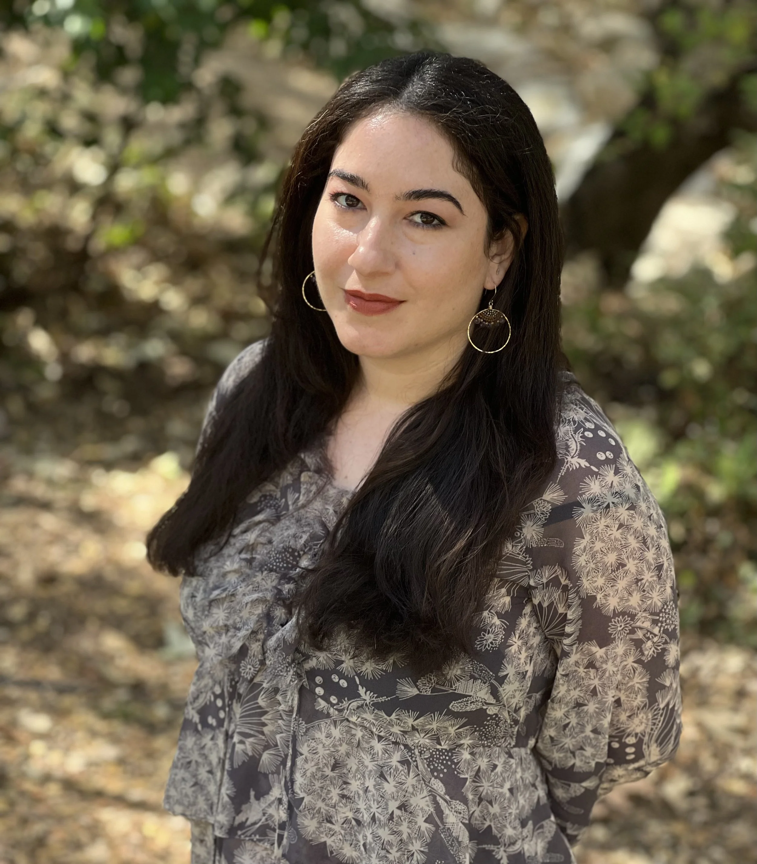 A woman with long dark hair and light skin standing outdoors among trees and blurred foliage, wearing a gray floral-patterned blouse and gold hoop earrings.