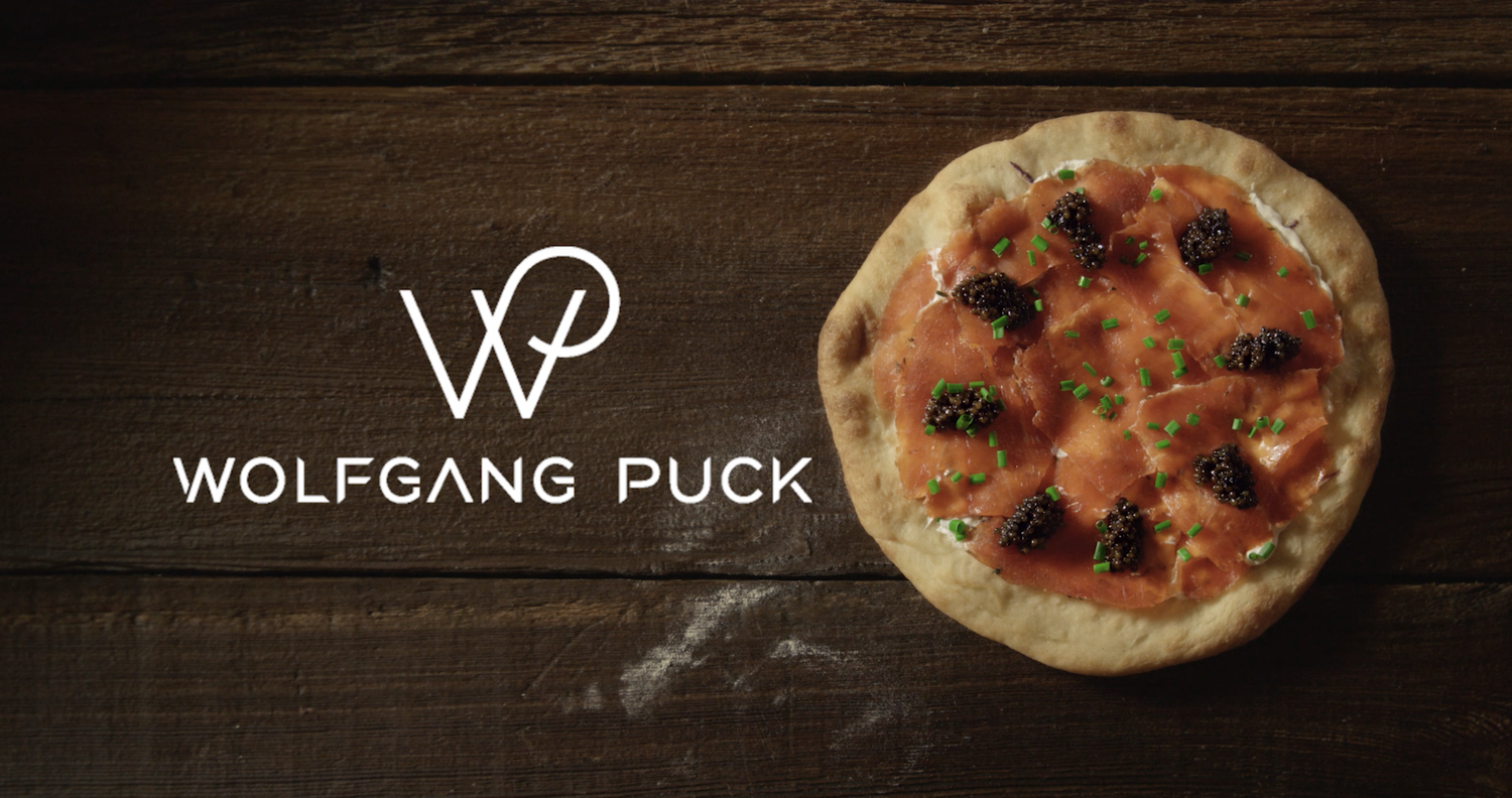 A pizza topped with smoked salmon, black caviar, and chopped chives on a wooden surface with Wolfgang Puck logo and text.