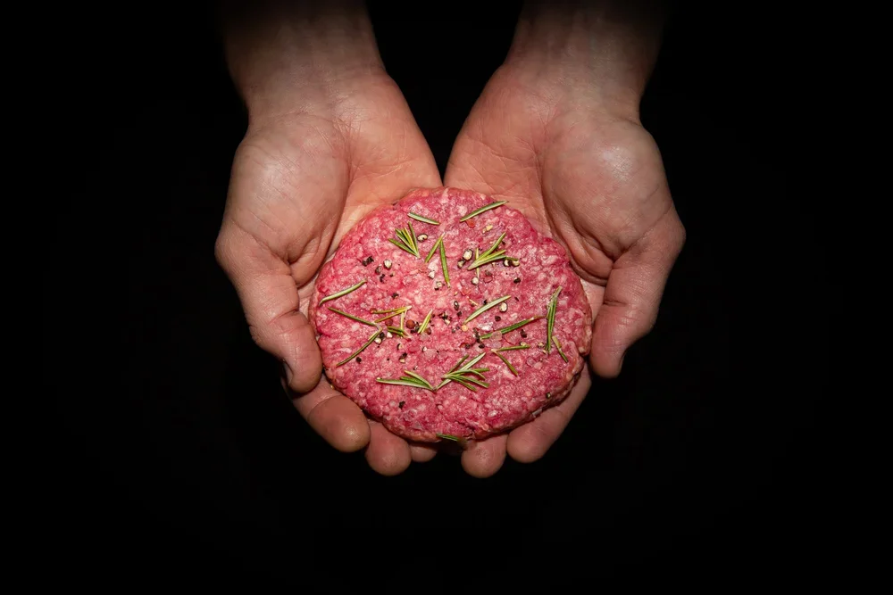 Hands holding a pink textured food item garnished with rosemary and black pepper on a black background.