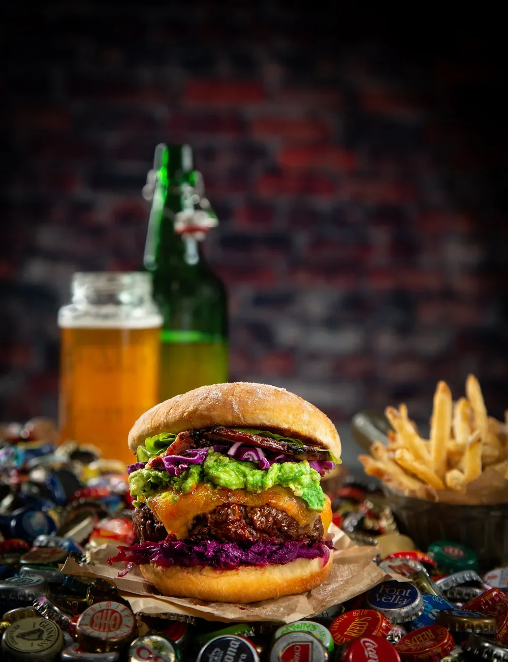 Cheeseburger with avocado, lettuce, red onions, and purple cabbage, with fries and beer bottles in the background on a table surrounded by bottle caps.