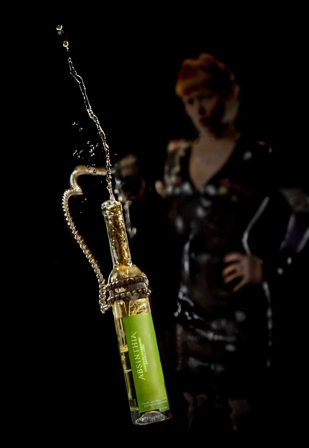 A bottle of Absinthiha alcohol is hanging from a chain, with liquid splashing out. In the background, a woman with red hair, wearing a patterned dress, is standing with her hands on her hips, blurred and out of focus.