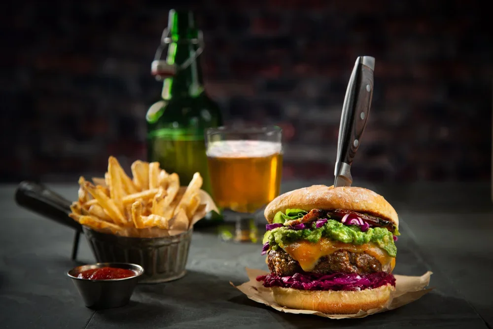 A cheeseburger with guacamole, lettuce, tomato, red cabbage, and melted cheese inside a toasted bun, with a knife stabbed upright through it, served with French fries, ketchup, and a glass of beer, against a dark backdrop.
