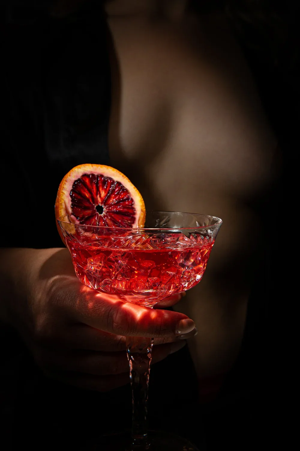 A hand holding a glass of red cocktail garnished with a blood orange slice.