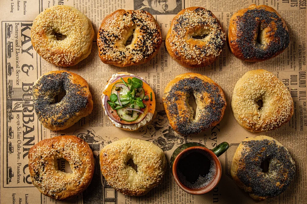 A tray of twelve bagels with various toppings, a small salad, and a mug of coffee on a newspaper.