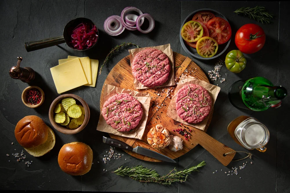 Raw hamburger patties with rosemary on a wood cutting board surrounded by ingredients for burgers including sliced cheese, pickles, tomato slices, onions, a bun, condiments, and seasonings on a dark surface.