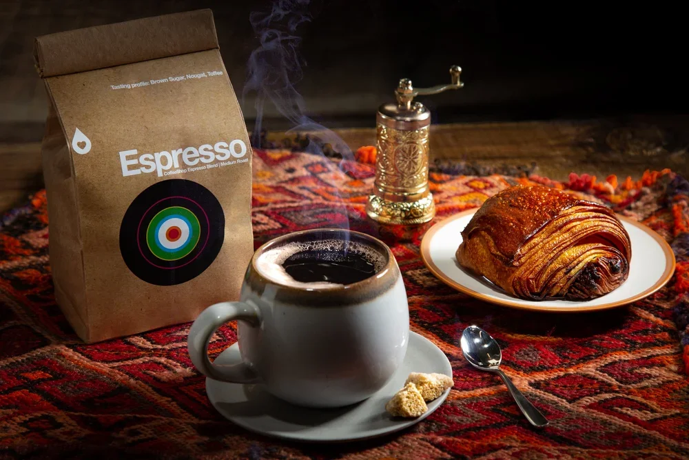 A steaming cup of black coffee on a saucer with sugar cubes, a plate with a chocolate croissant, a bag of espresso coffee beans, and a decorative pepper grinder on a colorful woven tablecloth.