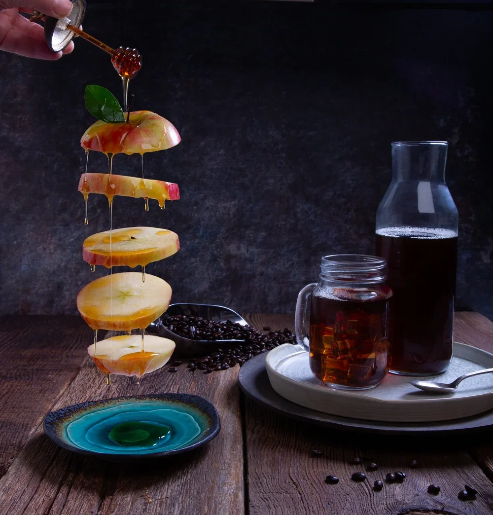 A tall skewer of apple slices drizzled with honey, with a honey dipper above applying more honey, set on a rustic wooden table.