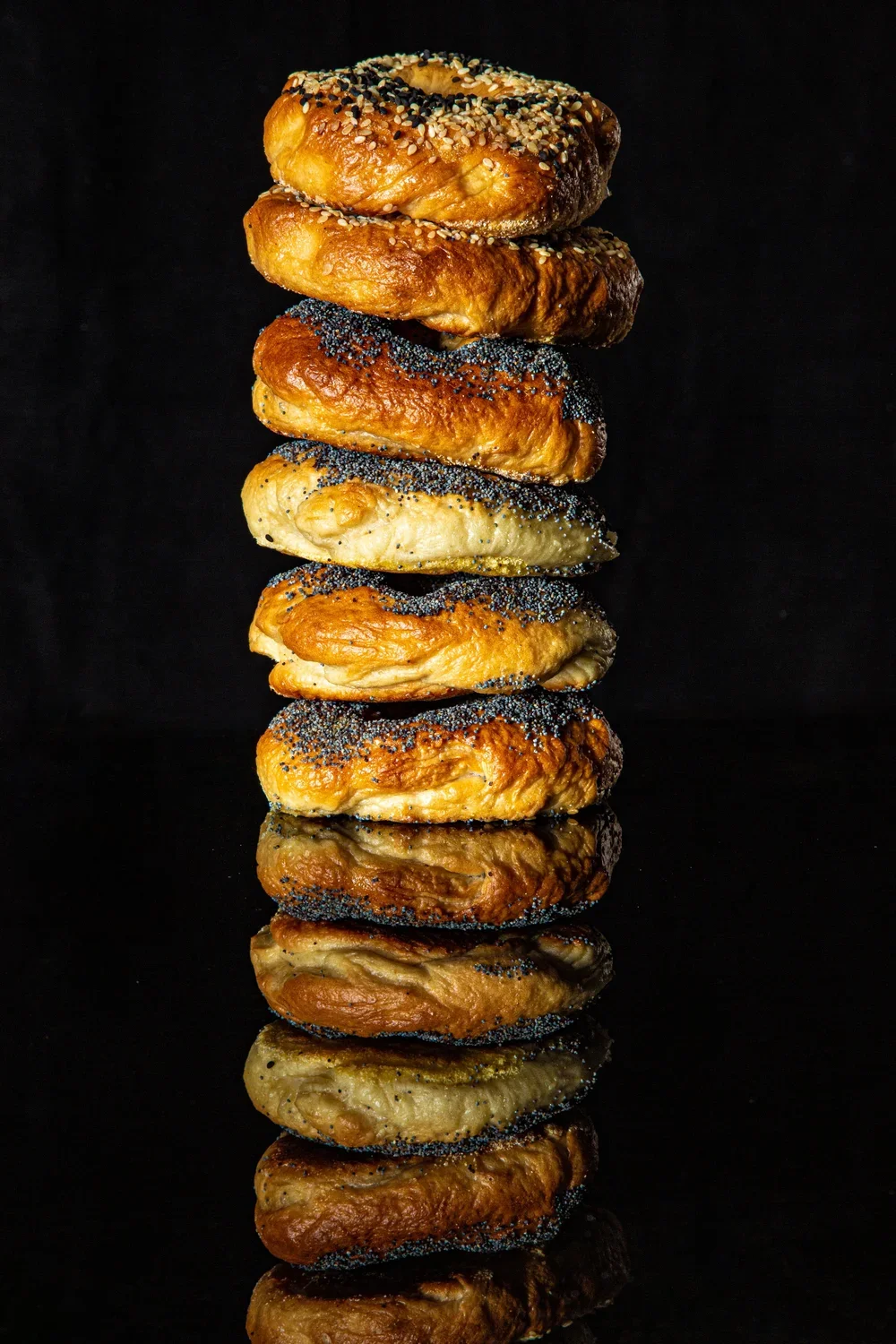 A tall stack of assorted, golden-brown bagels with sesame and poppy seeds on a black background with reflection.