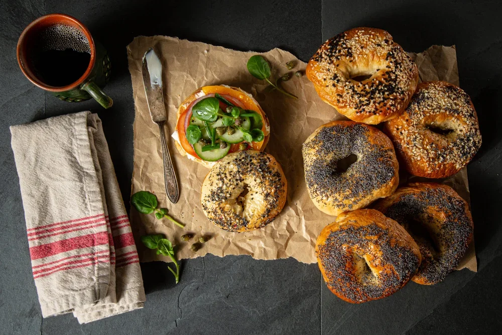 An assortment of bagels with poppy seed, sesame seed, and multigrain toppings, some plain, laid out on brown parchment paper. A bagel sandwich with cucumber slices, tomato, and greens is positioned among the bagels. A knife rests on the parchment, an